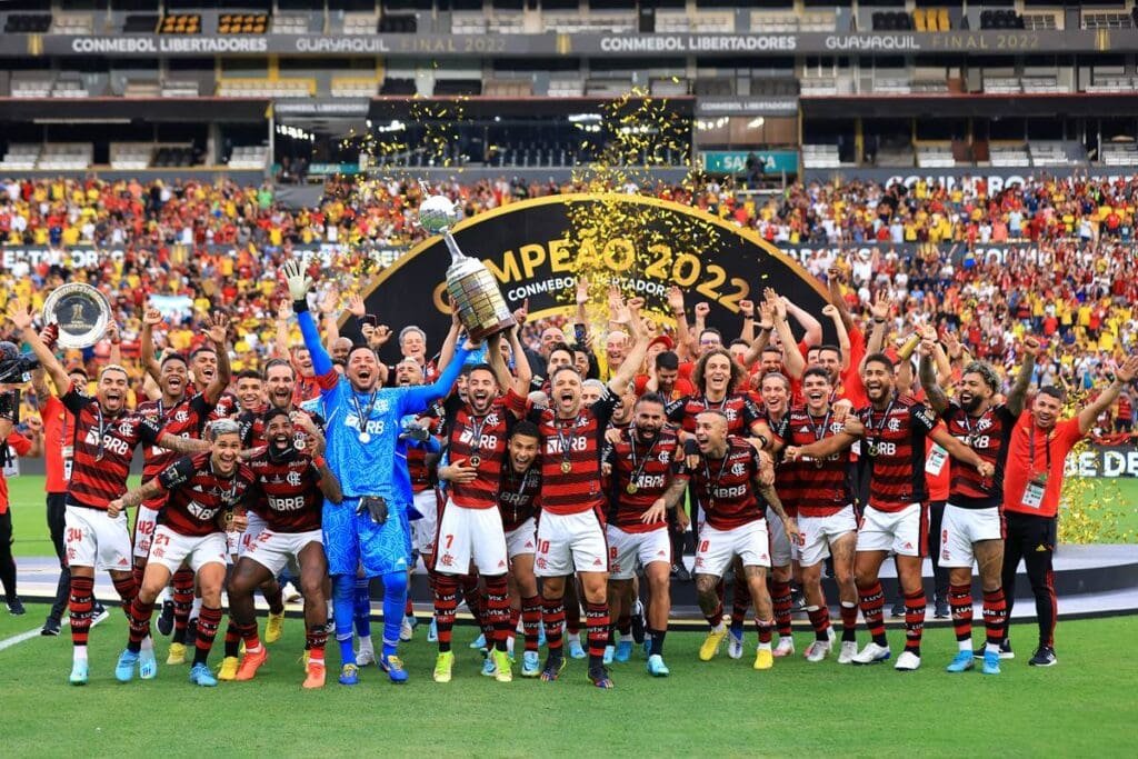 Elenco do Flamengo levanta troféu de campeão da Libertadores 2022 com chuva de confetes dourados e totem ao fundo escrito "Campeão 2022"