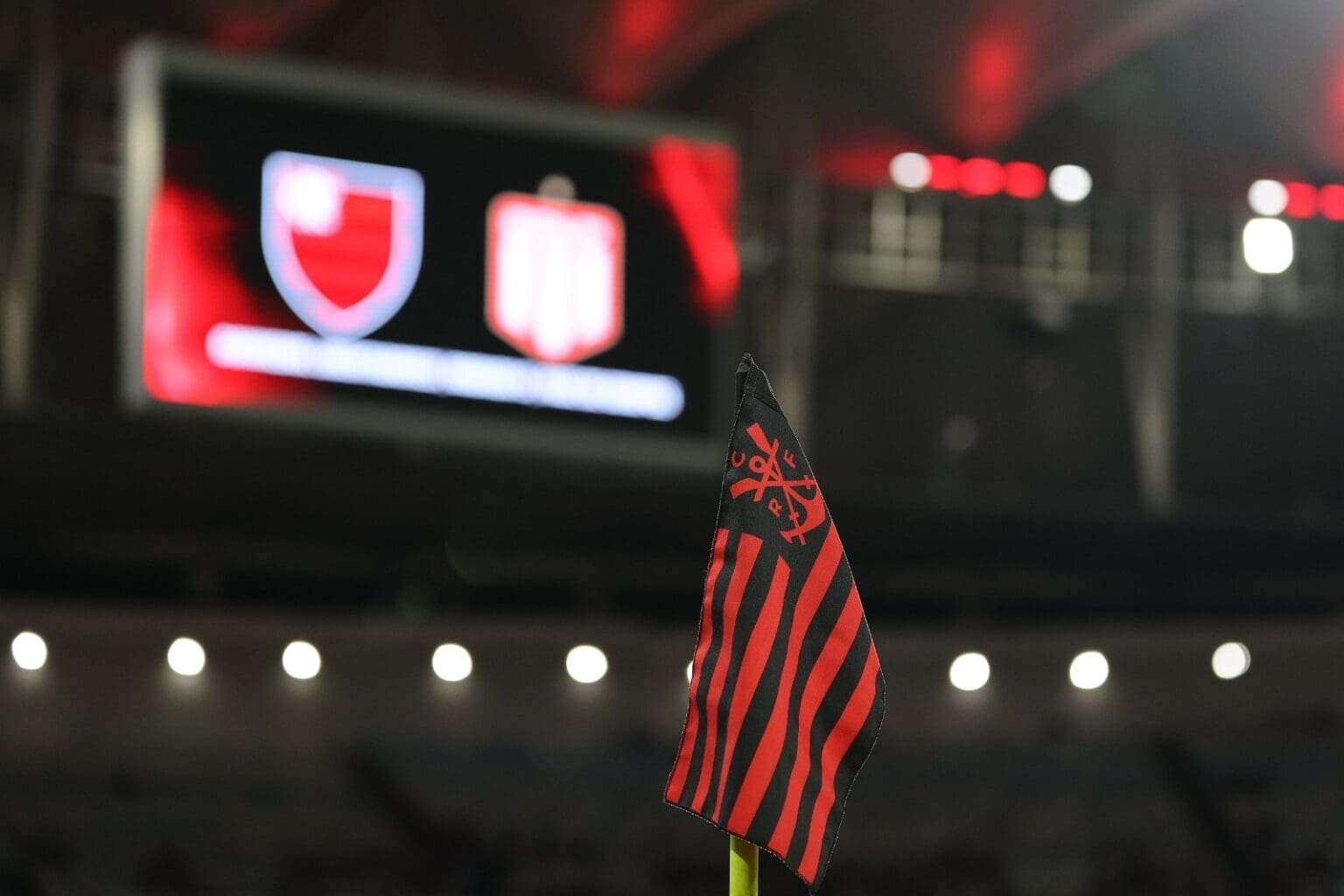 Conmebol denuncia Flamengo por infrações contra Estudiantes; confira penas Bandeira do Flamengo e, ao fundo, telão mostrando escudos de Flamengo e Estudiantes