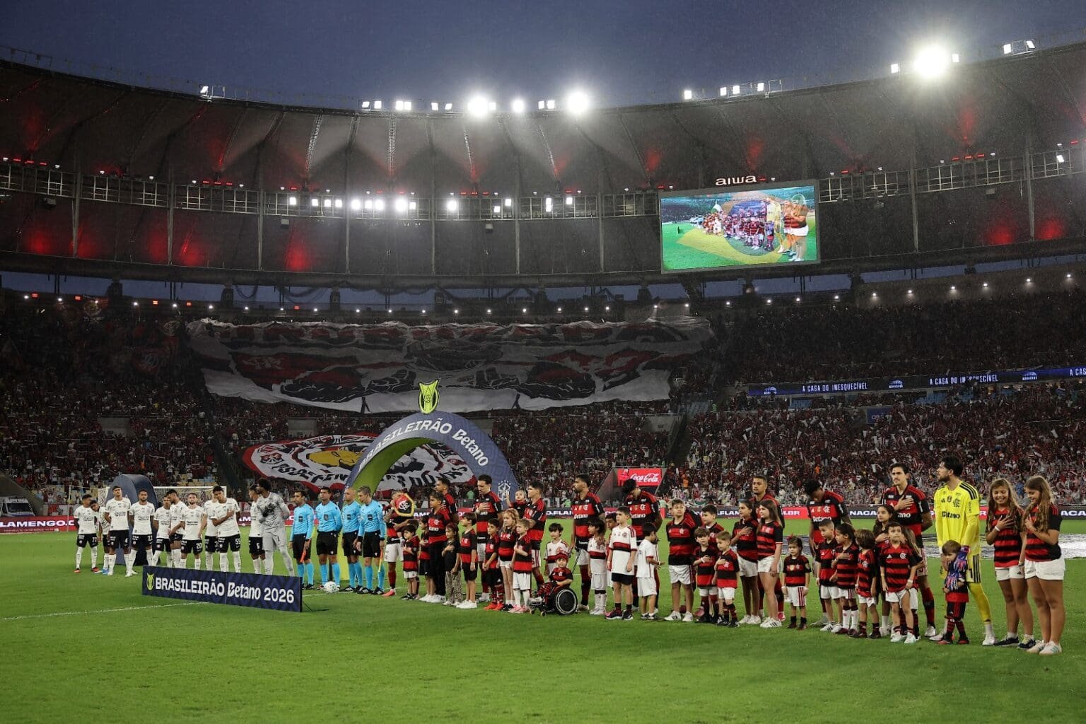 Jogadores de Corinthians e Flamengo perfilados para o hino nacional; ao fundo, bandeirão da urubuzada na arquibancada do Maracanã