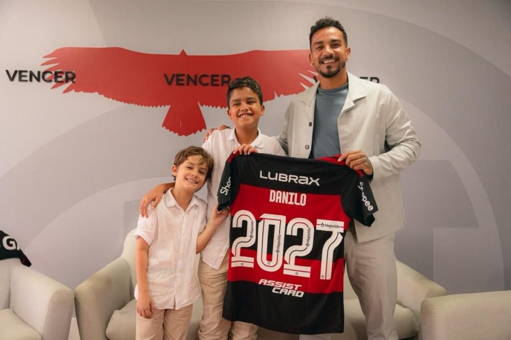 Flamengo oficializa renovação de Danilo até dezembro de 2027 Danilo sorri ao lado dos filhos ao posar com a camisa do Flamengo com o número 2027, ano do novo contrato